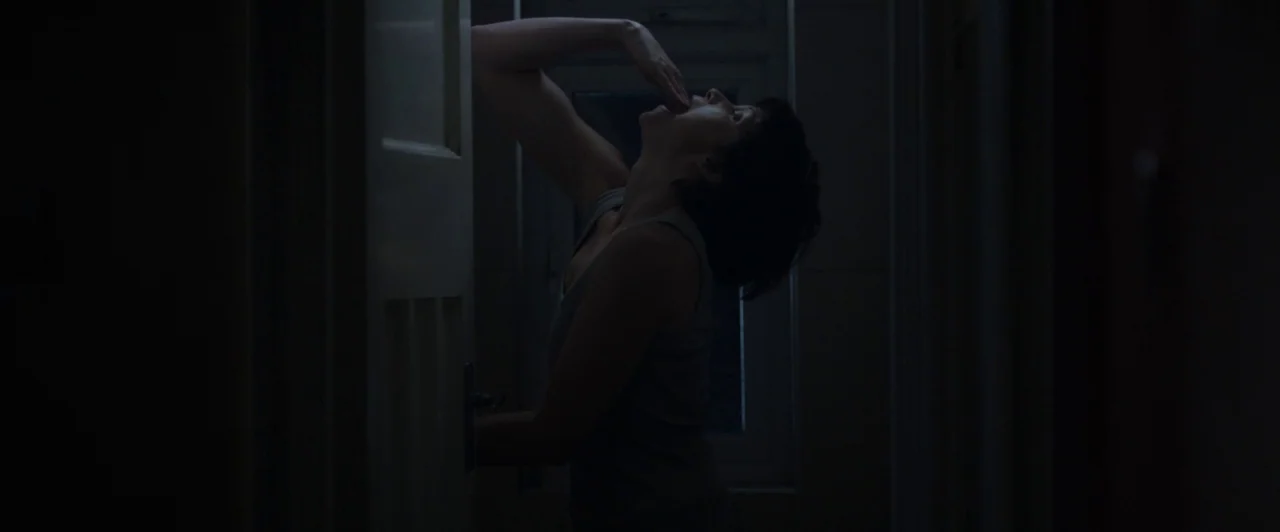 You Are Not My Mother (2021) movie still: medium — A dark, narrow hallway is lit only by dim light from a glass window. A woman stands in profile,…