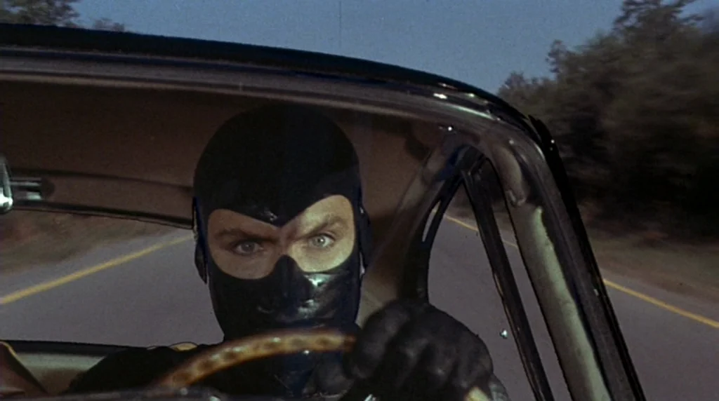 Danger Diabolik (1968) movie still: closeup — The interior of a moving car shows a driver wearing a black mask. The road and trees are visible…