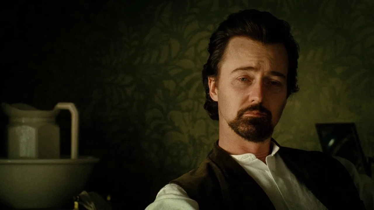 The Illusionist (2006) movie still: medium — A dimly lit washroom features dark green wallpaper with a faint floral pattern. A man in a vest sits…