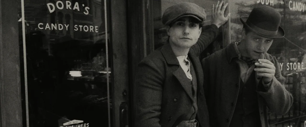 The Alto Knights (2025) movie still: medium — A vintage store exterior features a glass window with painted white lettering. Two men in period hats…