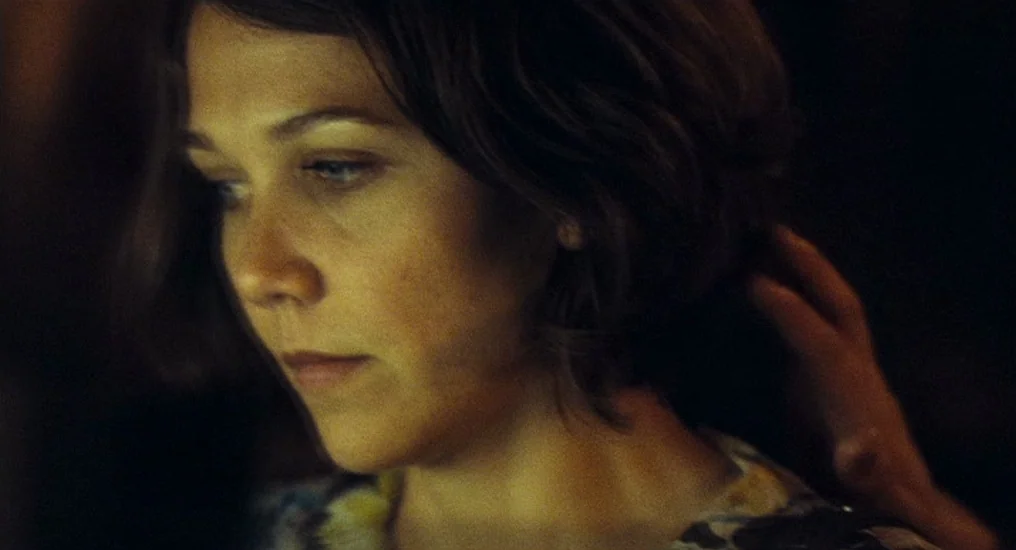 Paris, Je T’Aime (2006) movie still: closeup — A dimly lit interior space with a dark background. A woman with short hair looks down as a hand…