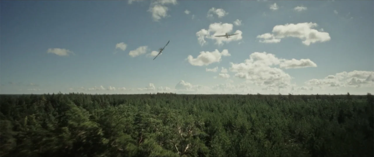 Sisu: Road to Revenge (2025) movie still: wide — Two propeller planes fly above a sprawling expanse of dense green pine forest. The aircraft are…