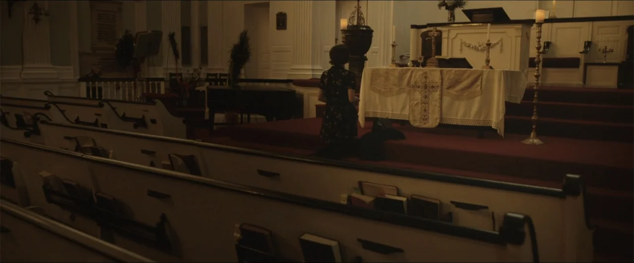 Exposed (2016) movie still: medium — An interior church altar area with wooden pews in the foreground and red carpeting. A woman kneels in a dark…