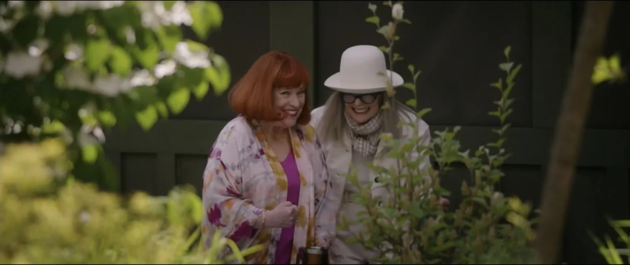 Summer Camp (2024) movie still: medium — A green outdoor wall stands behind two women surrounded by out-of-focus foliage. The women are standing…
