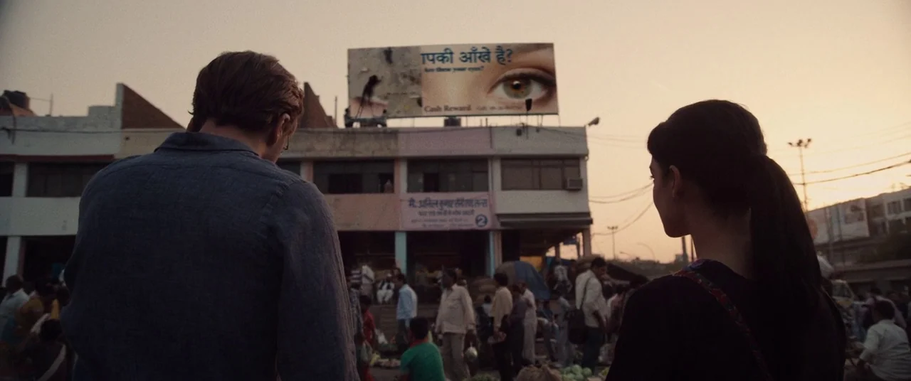 I Origins (2014) movie still: wide — A crowded Indian marketplace at sunset is overlooked by a large eye advertisement billboard. A man and woman…