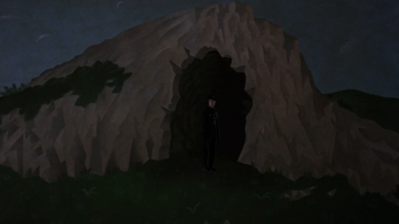 The Lair of the White Worm (1988) movie still: wide — A dark rocky landscape features a large crag with a deep central cavity. A man in a dark…