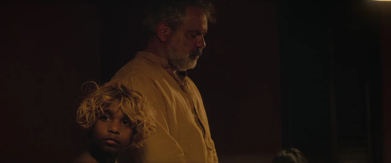 The New Boy (2023) movie still: medium — A dimly lit room with dark surroundings hides the architectural details from view. An elderly man and a…