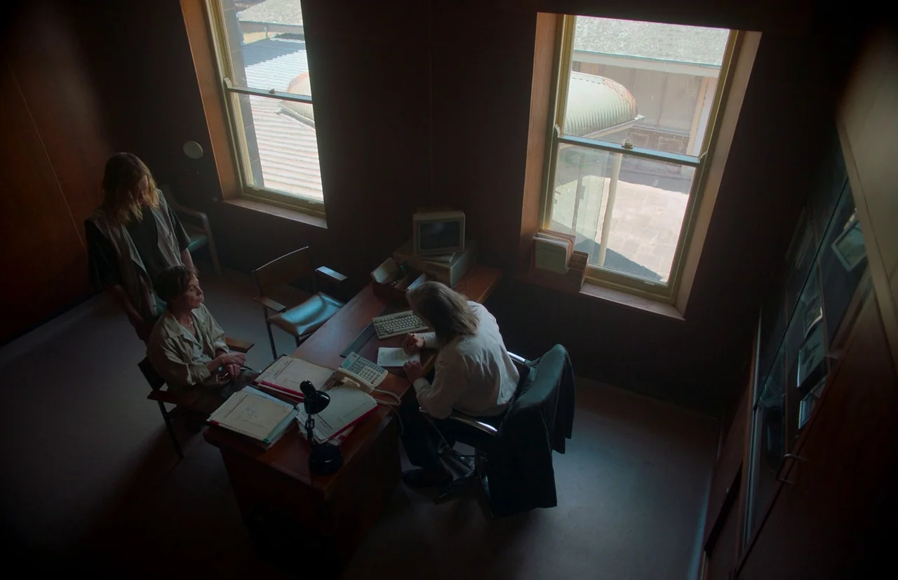 Nitram (2021) movie still: wide — A wood-paneled room features a desk cluttered with documents and an old computer monitor. A person writes at the…