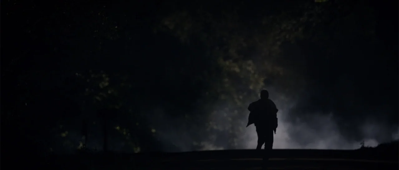 Jeepers Creepers 3 (2017) movie still: fullbody — A dark, unlit road at night features dense fog rolling across the ground surface. A lone figure in…