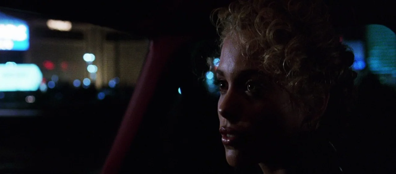 Showgirls (1995) movie still: closeup — A woman sits inside a car at night near a brightly lit urban area. Neon city lights blur in the background…