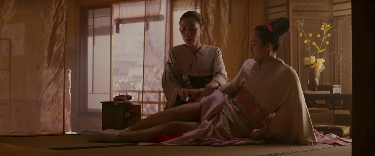 Memoirs of a Geisha (2005) movie still: medium — Two women wearing traditional kimonos sit on the tatami mats of a wooden house. One woman assists…