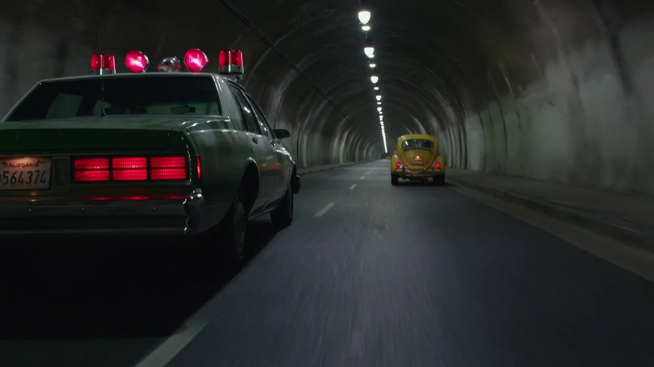 Bumblebee (2018) movie still: wide — A long, curved concrete road tunnel lit by evenly spaced overhead fluorescent fixtures. A green sedan drives in…