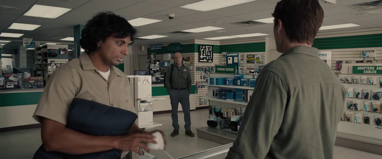 Glass (2019) movie still: medium — An electronics store interior with white shelving units displaying various cables and storage devices. Two…