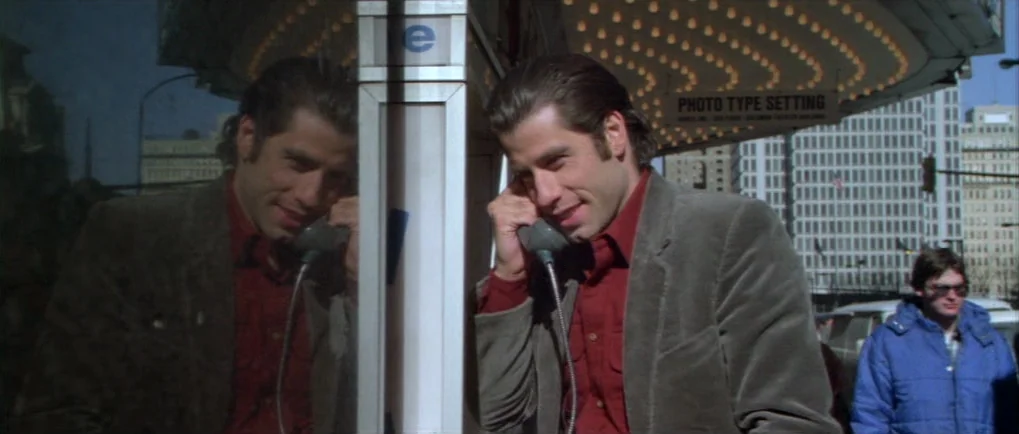 Blow Out (1981) movie still: medium — A man stands inside a glass-walled phone booth with a city street visible behind him. He smiles while holding…