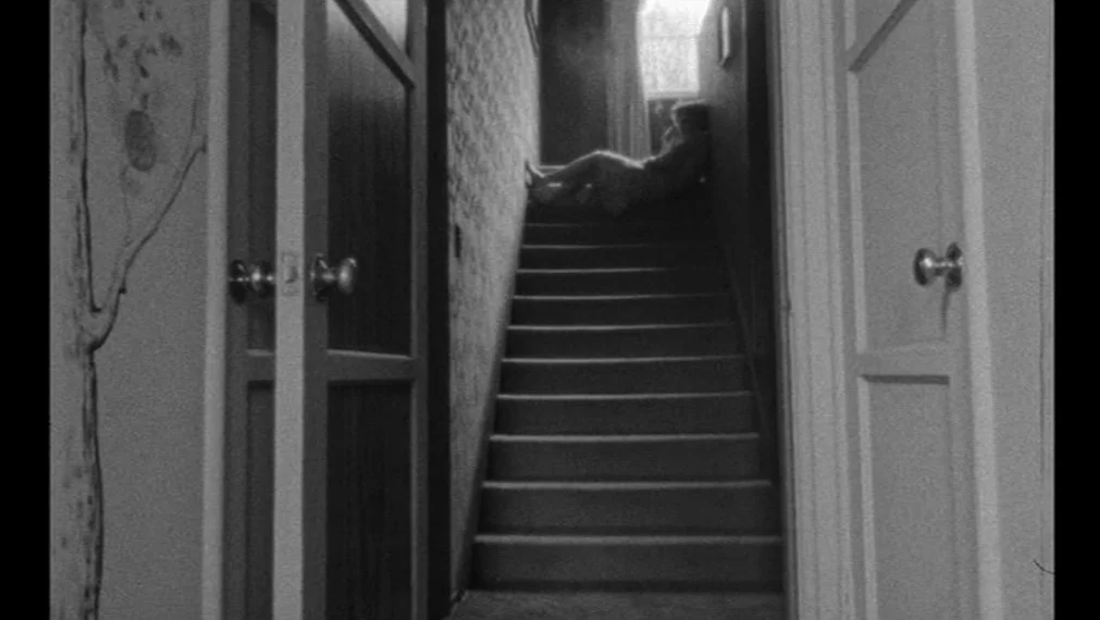 Faces (1968) movie still: wide — A dark, narrow residential staircase viewed from a foreground doorway. A single person sits at the very top against…