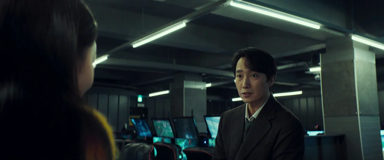 Decision To Leave (2022) movie still: medium — An interior shot of an internet cafe with rows of computer terminals and workstations. A man in a…