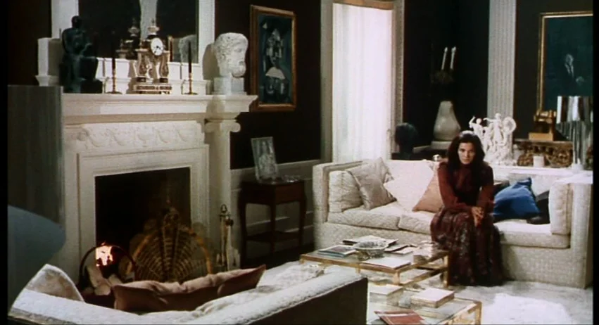 A Lizard In A Woman’s Skin (1971) movie still: wide — A dark-walled room features a large fireplace with a mantle and paintings. A woman sits on a…