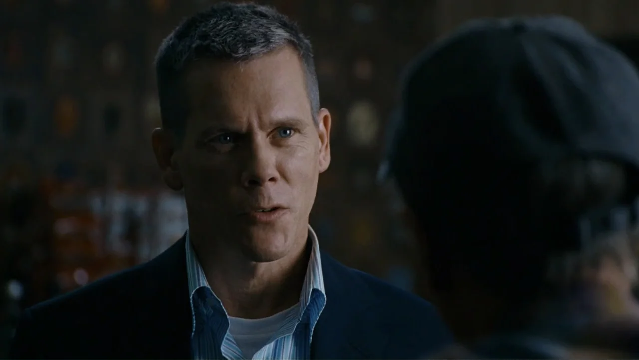 Taking Chances (2011) movie still: closeup — A man in a dark blazer talks to someone in a dark interior space. The background is dimly lit and…