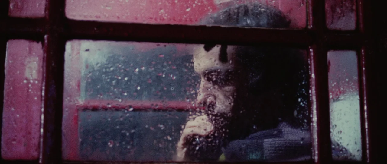 Death Walks On High Heels (1971) movie still: closeup — A man stands inside a classic red glass telephone box during a rainy night. He speaks on a…