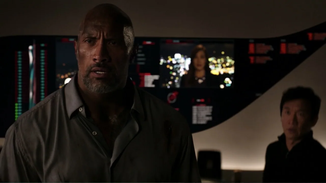 Skyscraper (2018) movie still: medium — A dimly lit high-tech control room features a curved display monitor showing a video feed. Two men stand in…