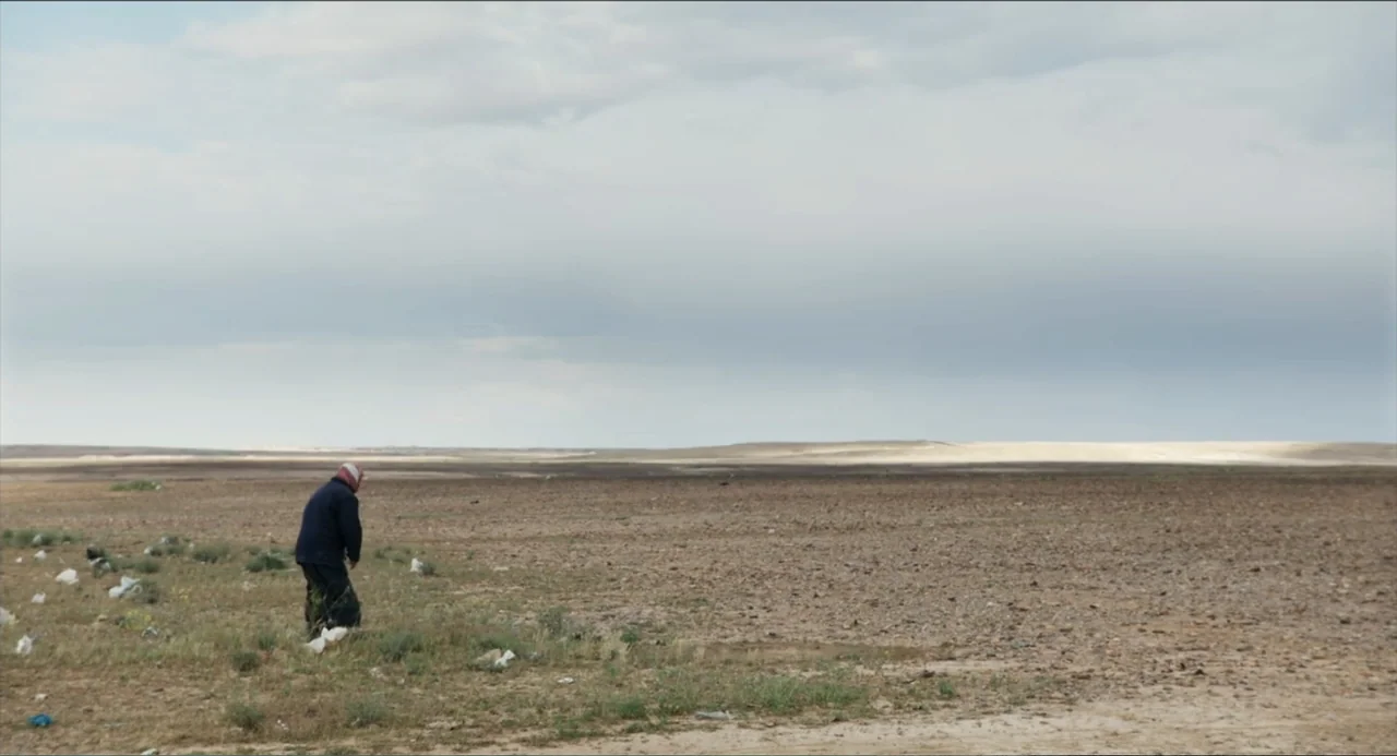Human Flow (2017) movie still: wide — An expansive arid desert landscape under a vast grey overcast sky. A person stands in the lower left, stooping…