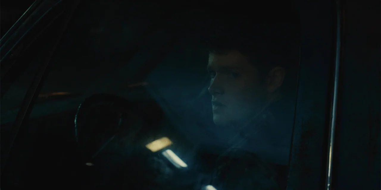 All Roads to Pearla (2019) movie still: closeup — The interior of a car at night is dark and dimly lit. A young man sits in the driver seat looking…