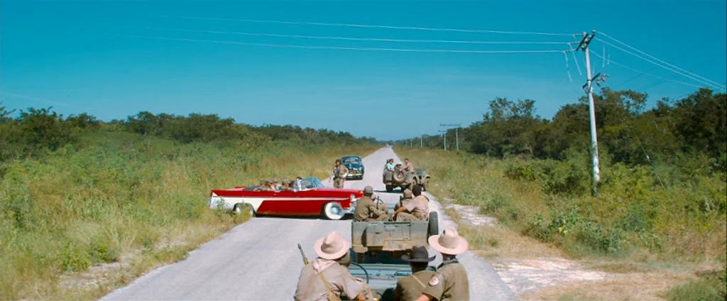Che Part 1 (The Argentine) (2008) movie still: wide — A flat asphalt road cuts through dense green foliage and scrub under a clear sky. Several…