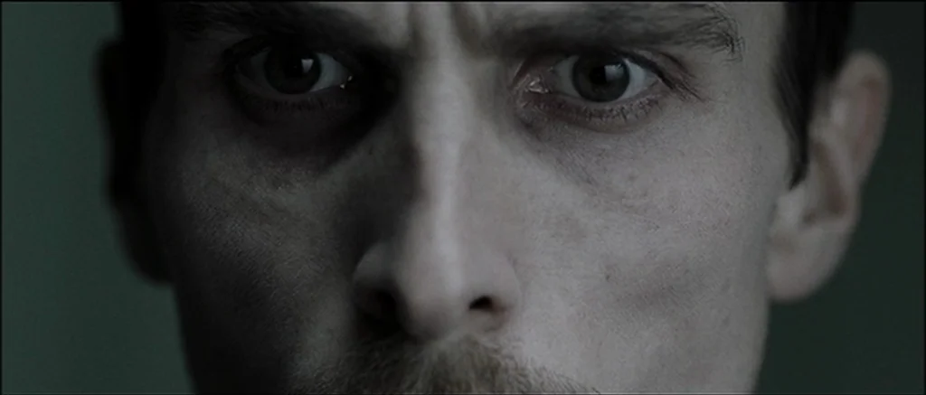 The Machinist (2004) movie still: closeup — A close-up view focuses on the eyes of a man against a dark background. The man maintains a steady gaze…