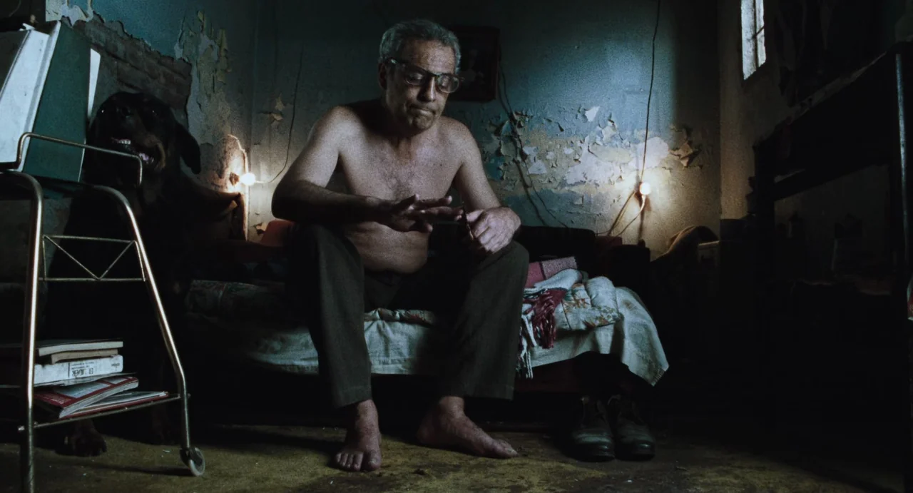Amores Perros (2000) movie still: medium — A dimly lit room features peeling paint on the walls and a cluttered bed. A shirtless man with glasses…