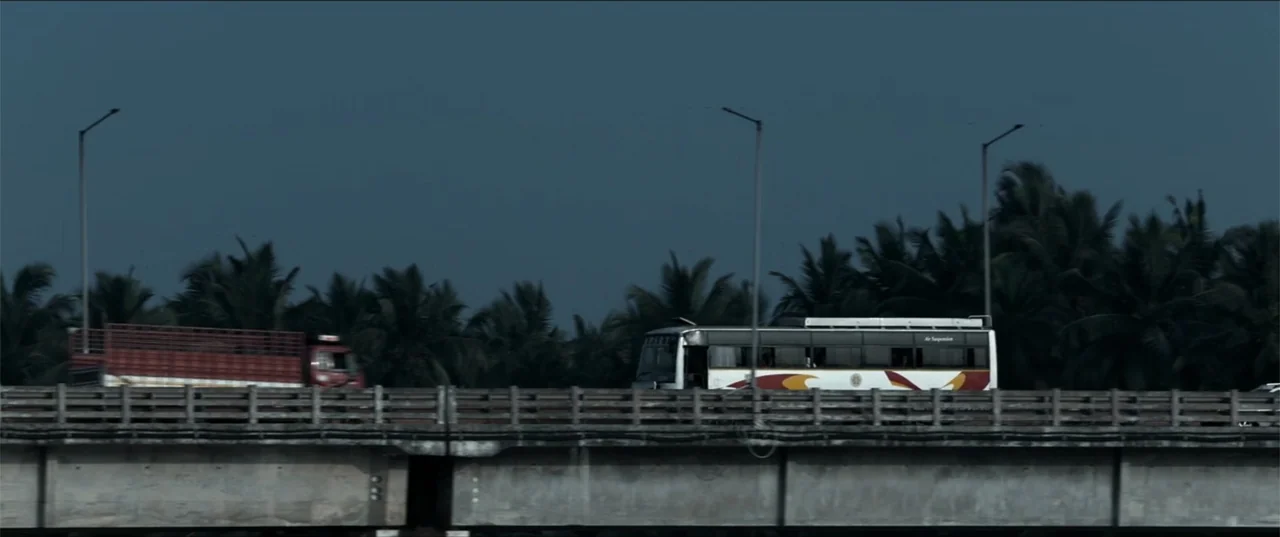 Cheekatilo (2026) movie still: wide — A concrete highway bridge spans a landscape filled with dark, silhouetted palm trees. A white passenger bus…
