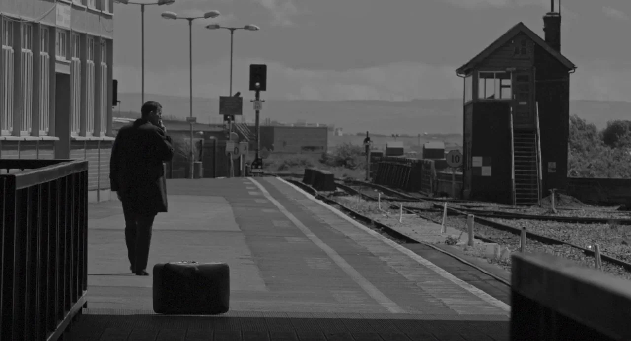 Song of Granite (2017) movie still: wide — A rural train station platform extends toward a small signal box in the distance. A man in an overcoat…