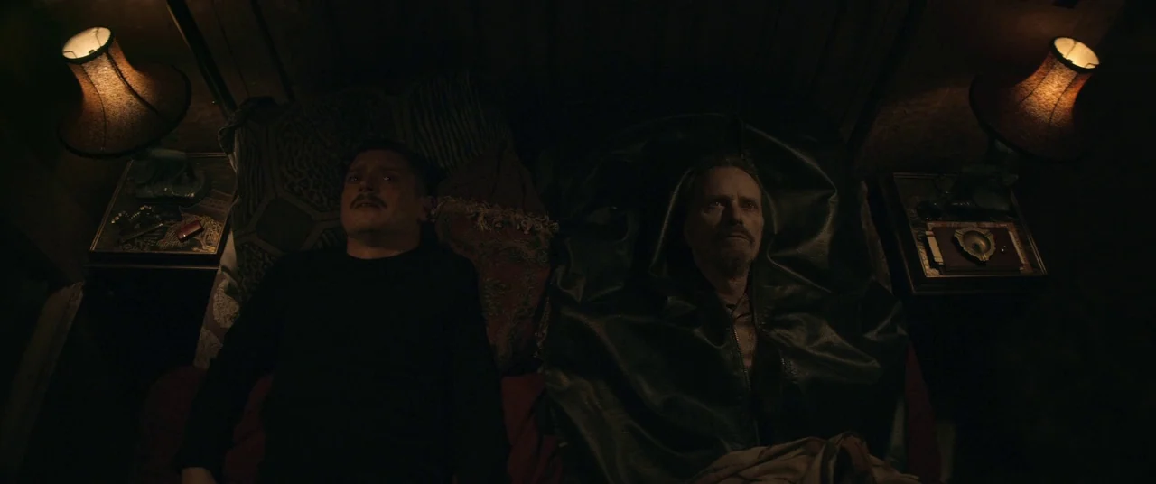 Come To Daddy (2019) movie still: medium — A dimly lit room with two men lying on a shared mattress at night. Each man has a glowing bedside lamp…
