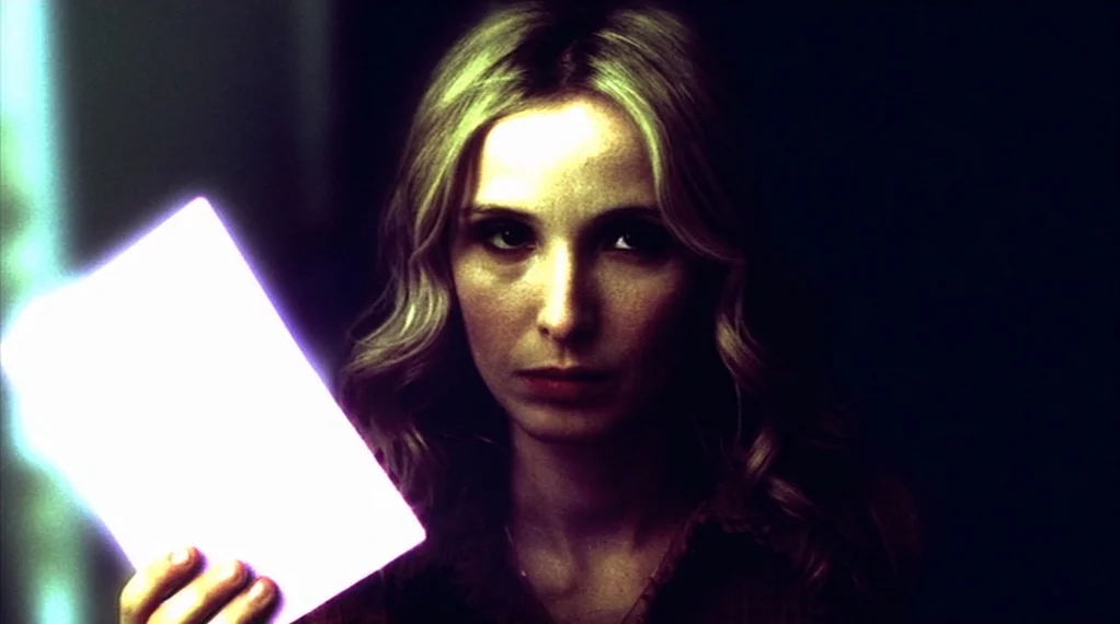 Broken Flowers movie still: closeup — A dimly lit interior space contains a woman holding an intensely bright rectangular object. The light from the…