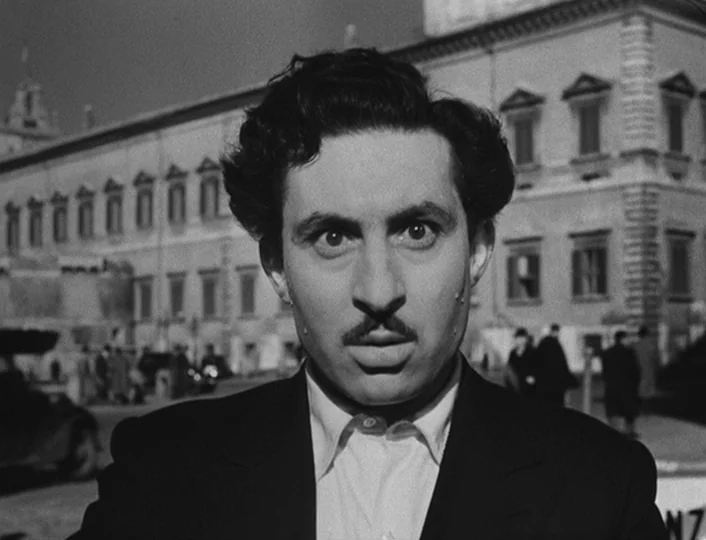 The White Sheik (1952) movie still: medium — A man stands in a city square in front of a classical stone building. He stares directly at the camera…