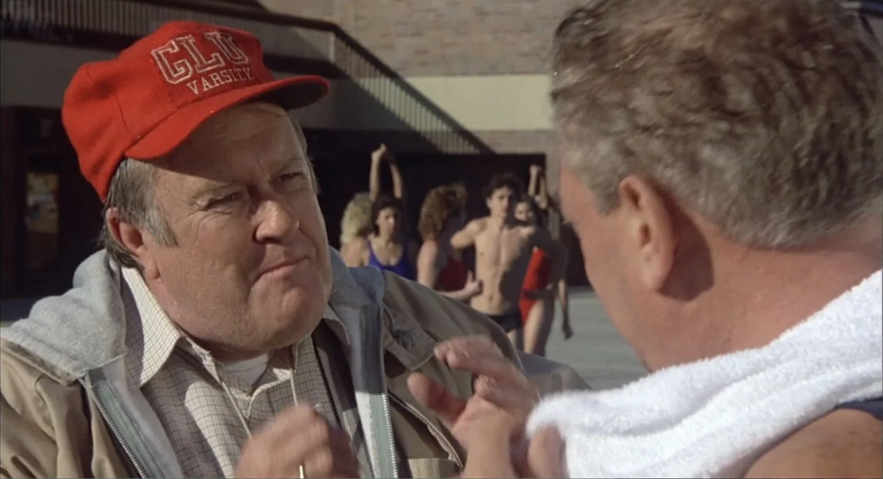 Back to School (1986) movie still: medium — A sunny outdoor pool deck with swimmers blurred in the background. Two middle-aged men converse while…