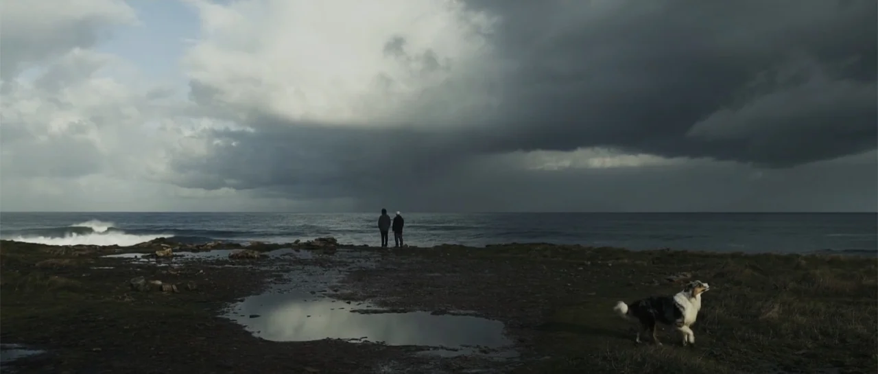 Taking Back the Waves (2017) movie still: wide — A rocky, windswept coastline under a dark, heavy overcast sky with rain pools. Two people stand…