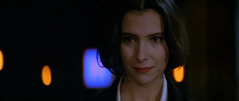 IP5 (1992) movie still: closeup — A dark, out-of-focus background features glowing orange and blue light sources. A woman with a dark bob haircut…