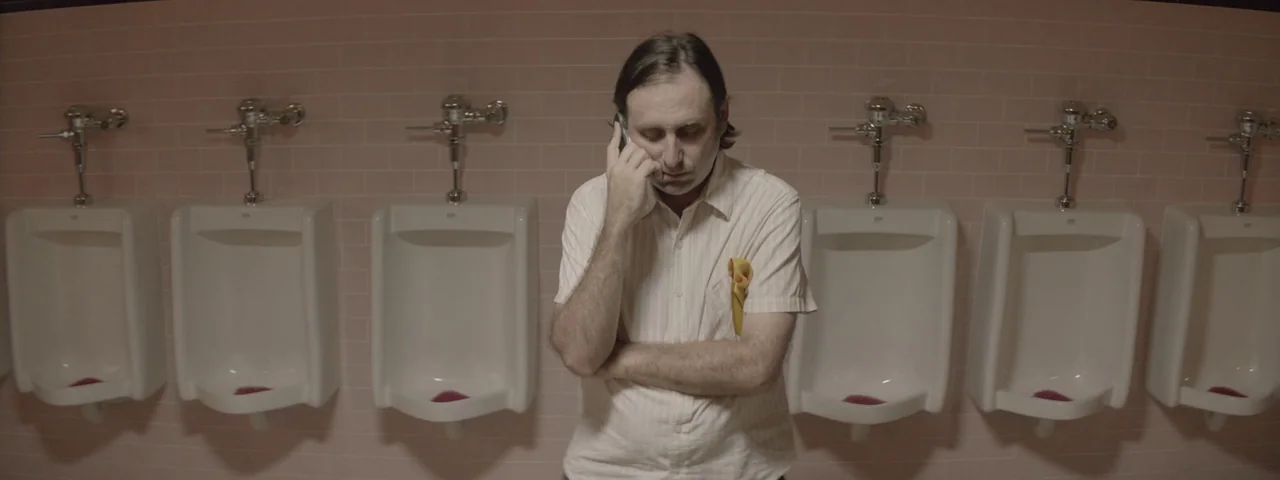 Entertainment (2015) movie still: medium — A row of white porcelain urinals is mounted against a wall of pink tiles. A man in a pinstriped shirt…