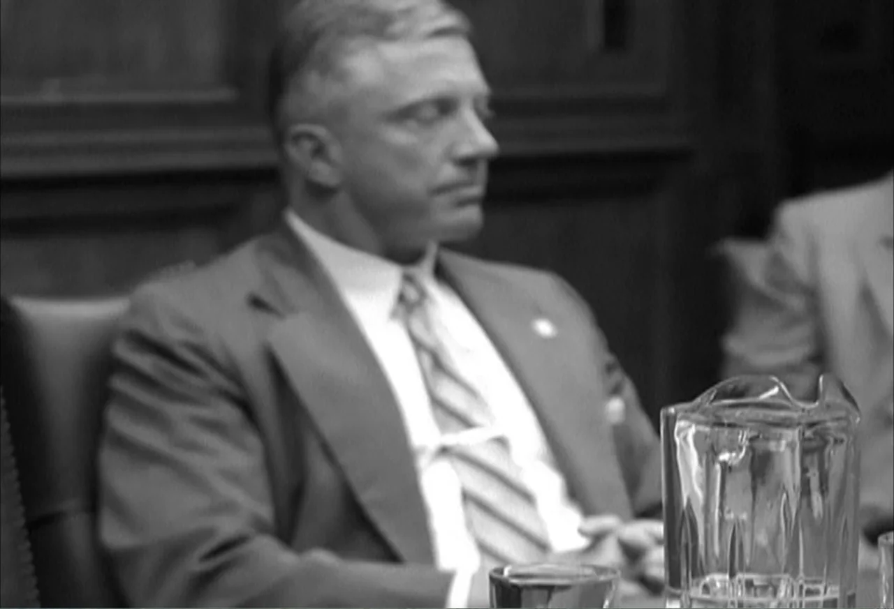 Boycott (2001) movie still: medium — A wood-paneled conference room features a dark table with a glass water pitcher. A man in a suit sits with…