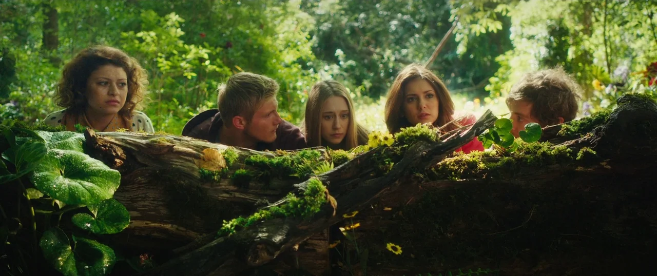 Final Girls (2015) movie still: medium — A dense green forest clearing with a large fallen tree trunk covered in moss. Five young adults crouch…