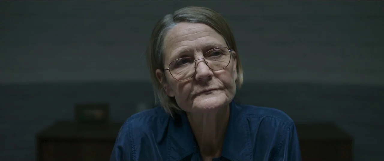 Border movie still: closeup — A dimly lit room with plain, dark gray walls provides a somber, sparse environment. An older woman with glasses wears…