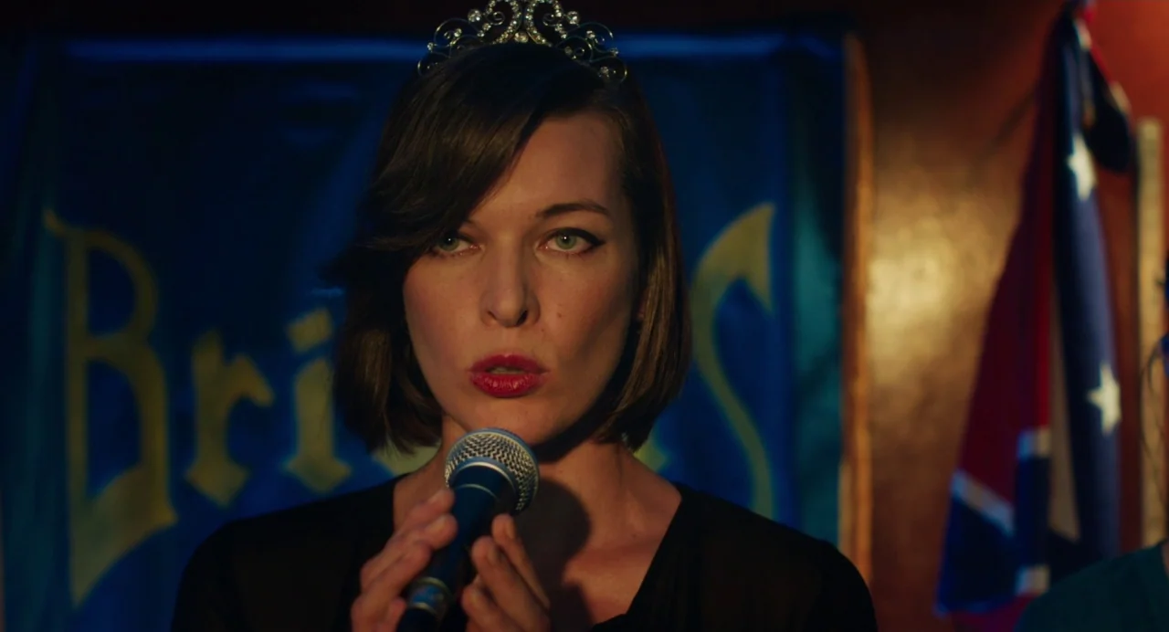 Cymbeline (2014) movie still: closeup — A woman stands in a dimly lit bar in front of a blue and gold banner. She speaks into a microphone while…