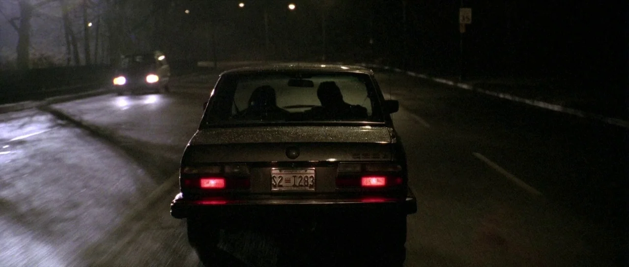 The Mothman Prophecies (2002) movie still: wide — A dark road at night is illuminated by car headlights and sparse overhead lighting. Two…