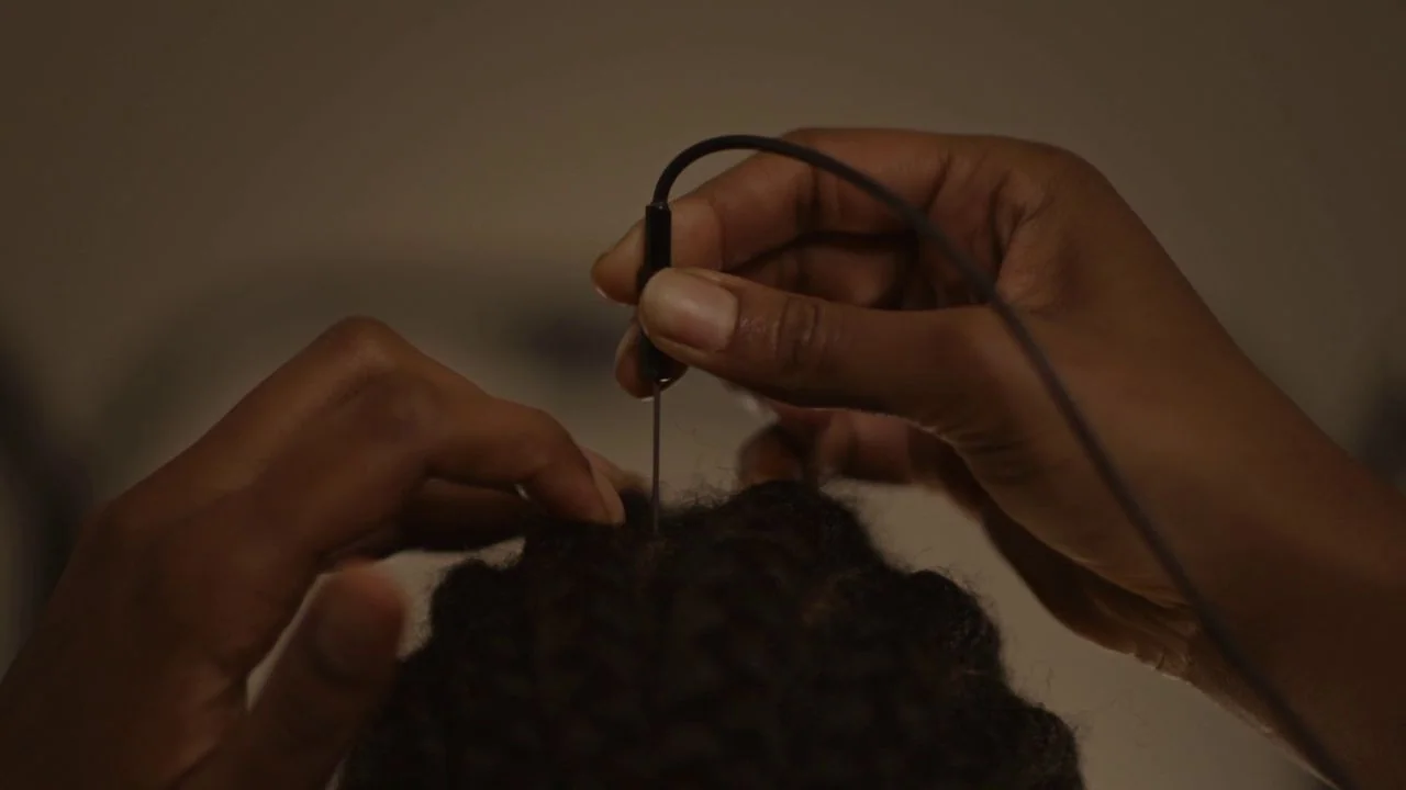 Possessor (2020) movie still: closeup — Hands work on a person's dark braided hair with a thin metal tool. The close focus obscures the surrounding…