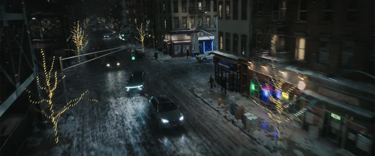 Play Dirty (2025) movie still: wide — A snowy urban intersection at night features brick buildings with storefronts and decorative lights. Two cars…