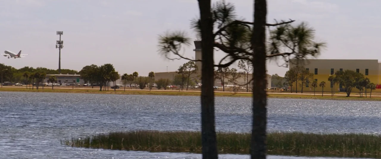 On a Wing and a Prayer (2023) movie still: wide — A flat lakeside landscape features airport facilities, a tower, and trees in the distance. A…