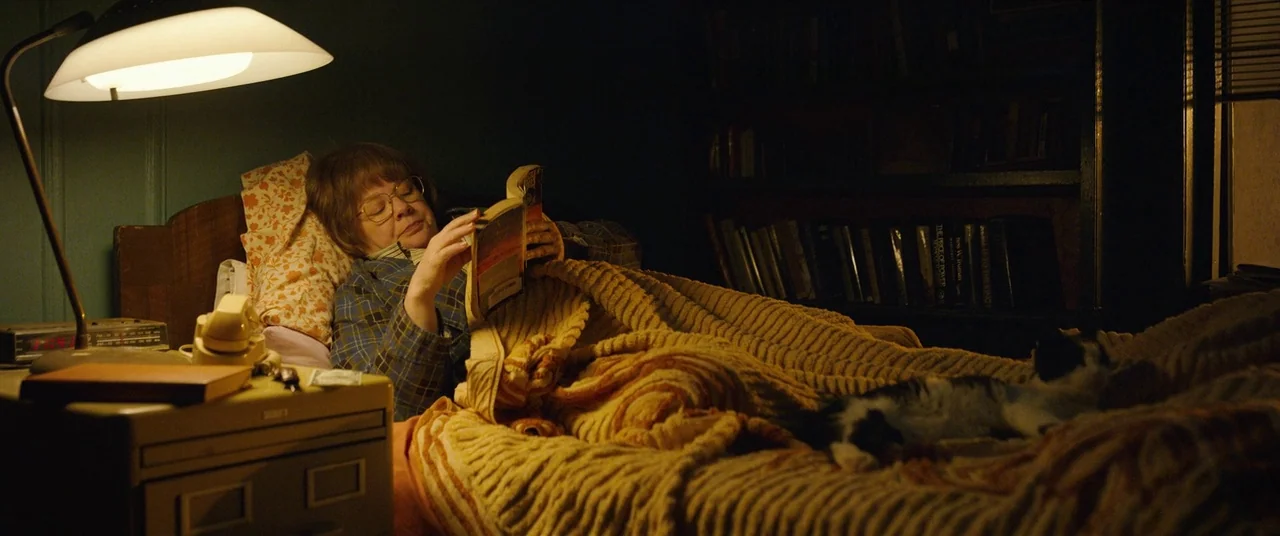 Can You Ever Forgive Me? (2018) movie still: medium — A darkened room contains a bed with a thick textured quilt and a bookshelf. An older woman…