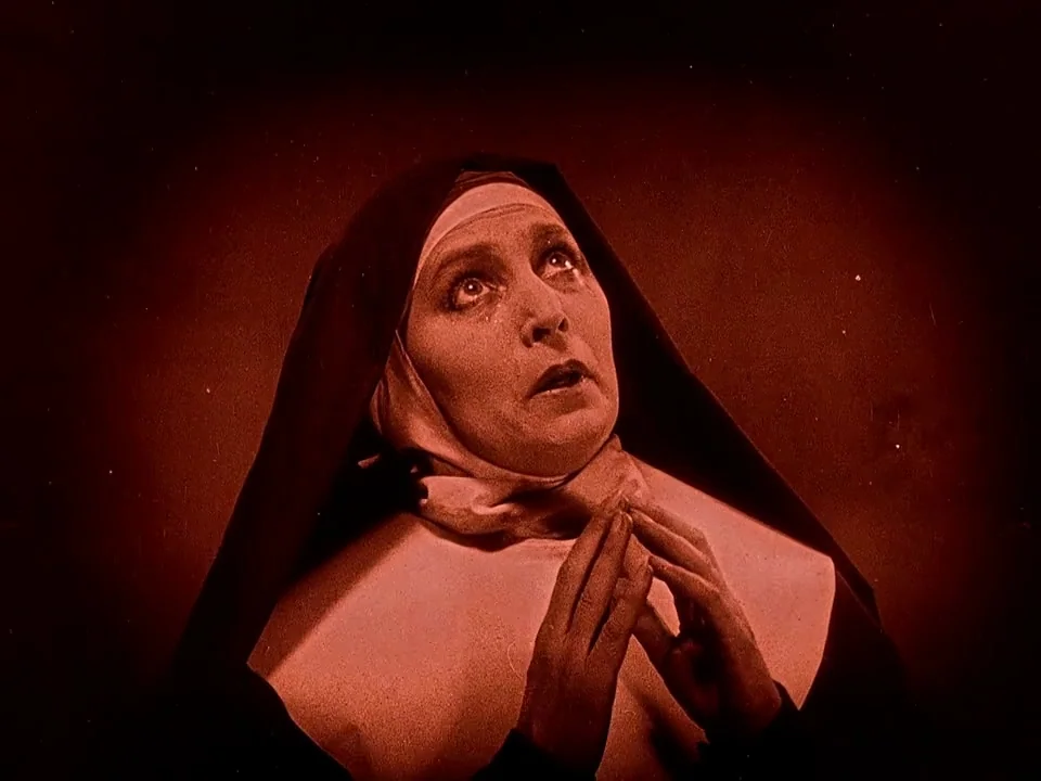 Häxan (1922) movie still: closeup — A woman dressed as a nun is positioned against a dark, undifferentiated background. She gazes upward with…
