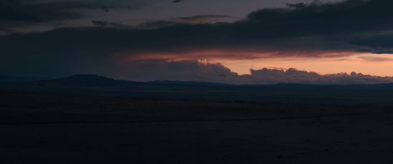 Intrusion (2021) movie still: establishing — A vast, dark, flat landscape extends toward distant rolling hills under a twilight sky. Thick storm…