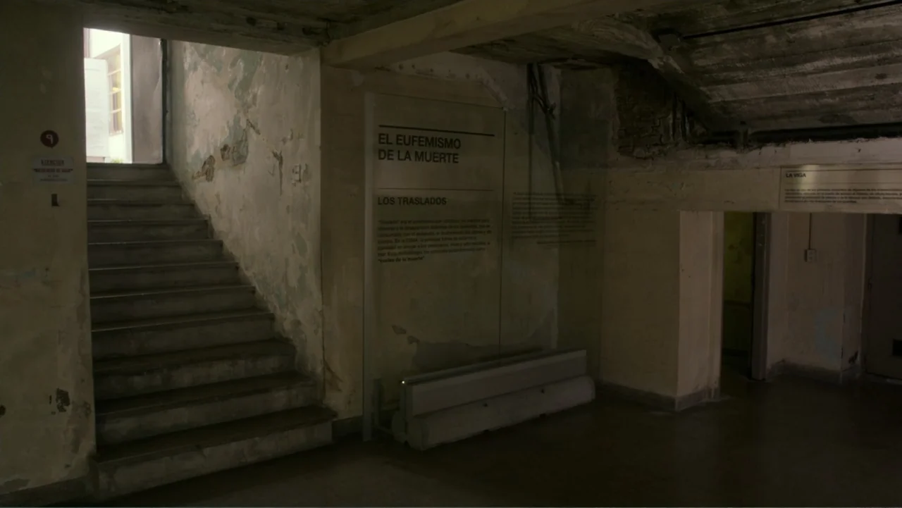 Transfers (2024) movie still: establishing — A dimly lit concrete stairwell with worn walls and informational wall plaques installed. The interior…